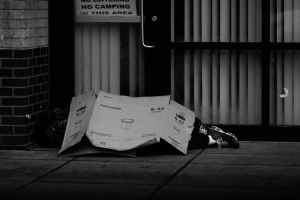 grayscale photo of human lying on ground covered of cardboard box